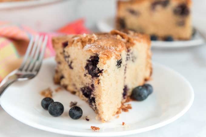 close up of slice of blueberry cake on white plate with fork and five whole blueberries