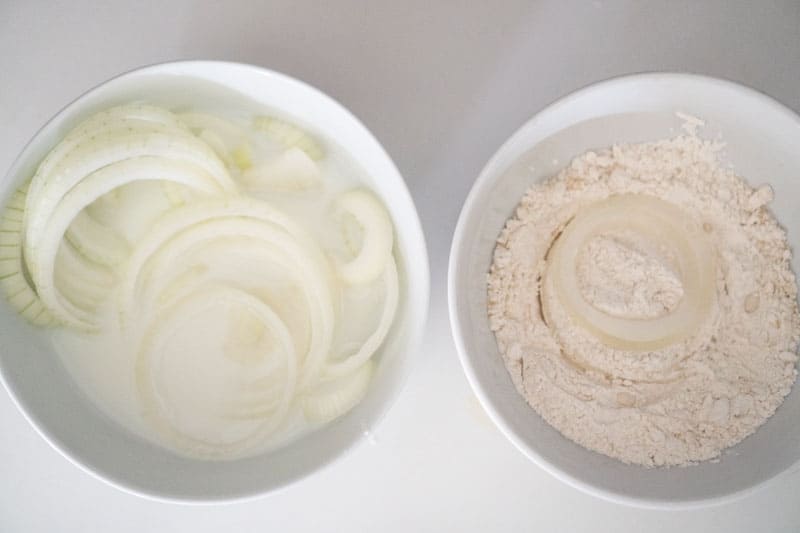 two white bowls one with milk and the other with flour both containing onion rings