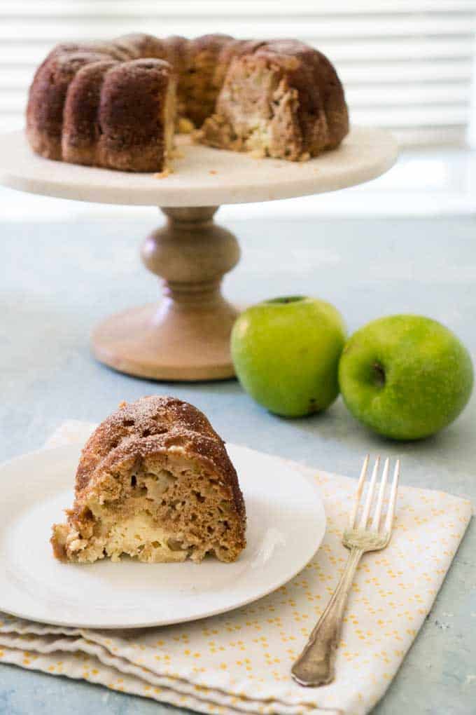 slice of apple cake with cake stand and two green apples in background