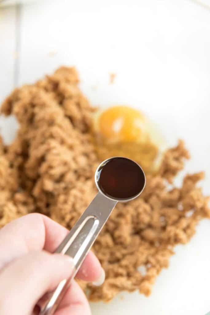 teaspoon of vanilla extract held over glass mixing bowl with dough and egg