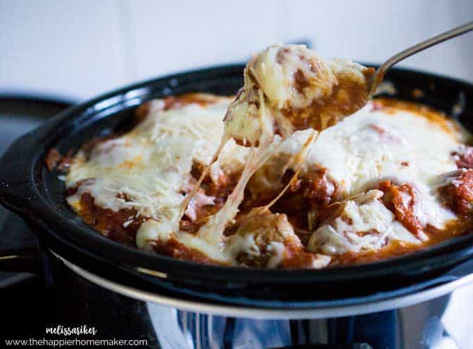 A slow cooker ravioli with meatballs topped with melted cheese with a spoon removing some ravioli 