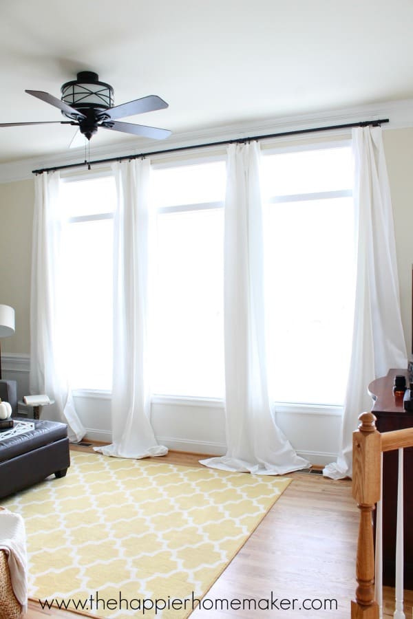 3 large windows with long white curtains hanging from dark curtain rod and yellow patterned rug on the wood floor