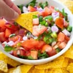 hand holding totrtilla chip with pico de gallo in white bowl in background