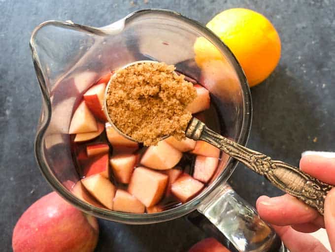 adding brown sugar to glass pitcher of sangria with chopped fruit