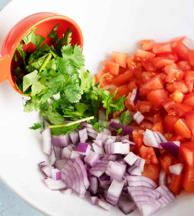 white bowl with chopped red onion, tomato and pouring cilantro our of measuring cup