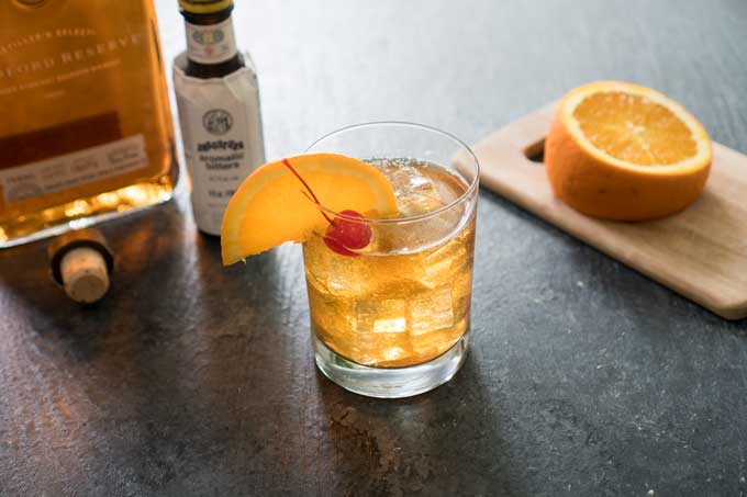 old fashioned bourbon cocktail with bottle of bitters and garnished with orange slice and cherry