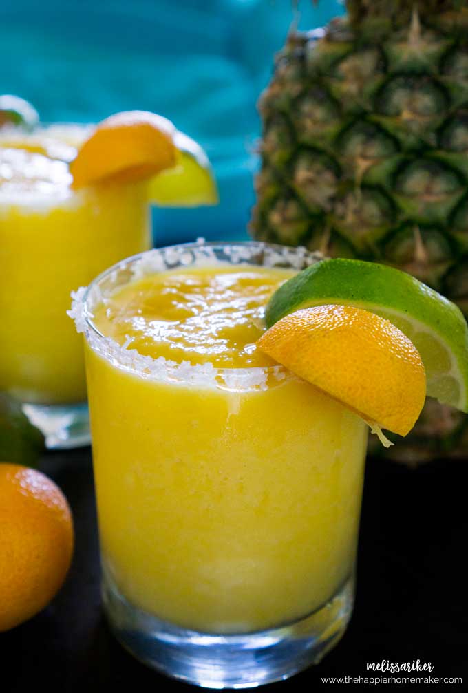 A close up of a frozen pineapple margarita garnished with a slice of lime and lemon in front of a pineapple