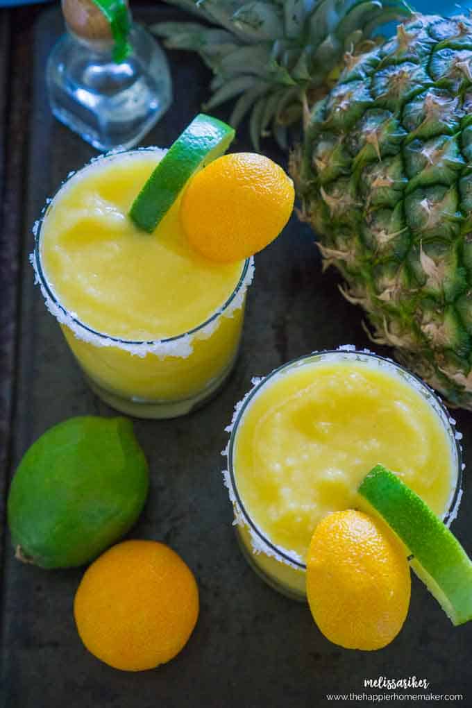 An above picture of two frozen pineapple margaritas garnished with a slice of lime and lemon in front of a pineapple