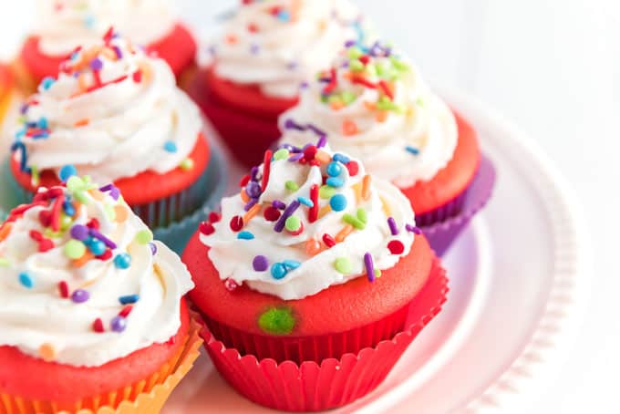A close up of a rainbow cupcake topped with white icing and sprinkles on white plate