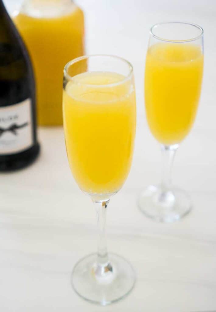 two mimosas in champagne flutes with prosecco bottle and orange juice in background on marble table